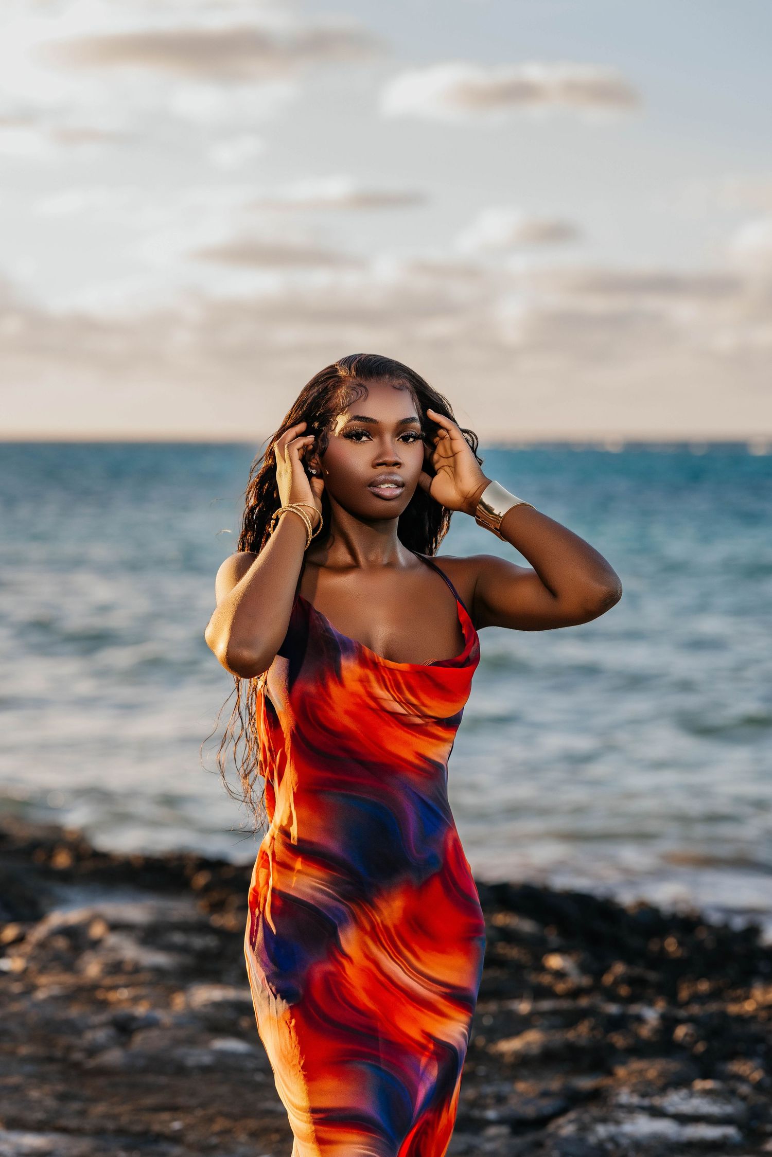 Woman in vibrant red and blue dress poses at sunset by ocean.
