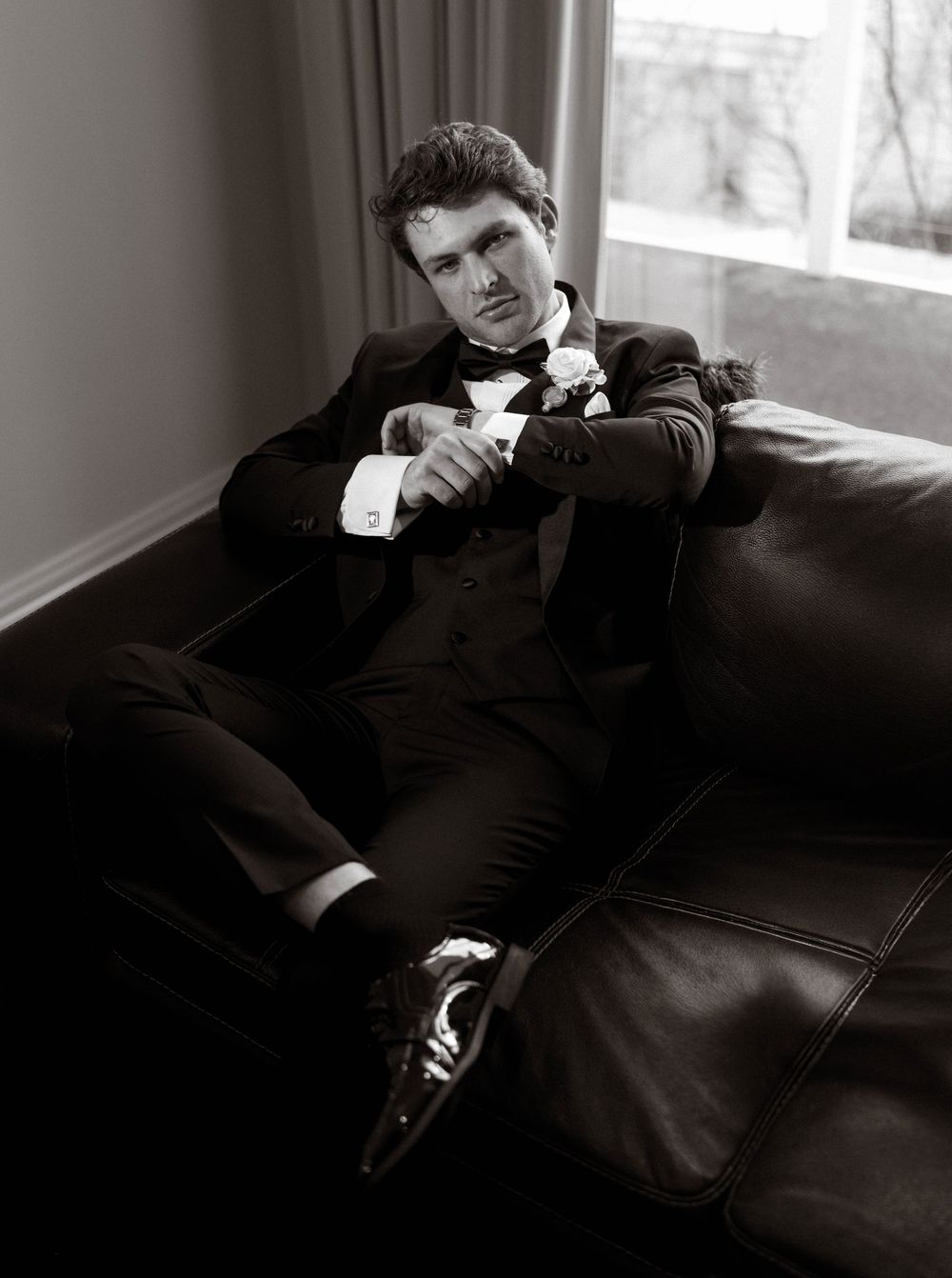 A person in black clothing lounges on a dark leather couch in dramatic black and white photography.