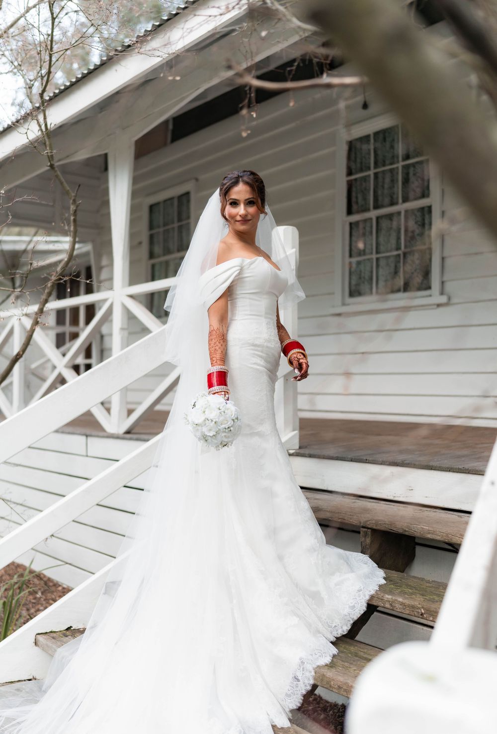 A bride in an elegant white lace gown with a flowing train poses near a rustic white porch.