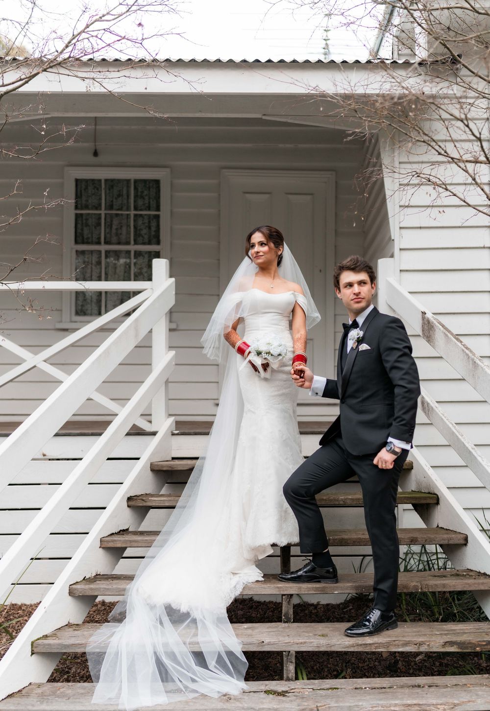 A newlywed couple sits together on white porch steps of a rustic farmhouse in a romantic moment.