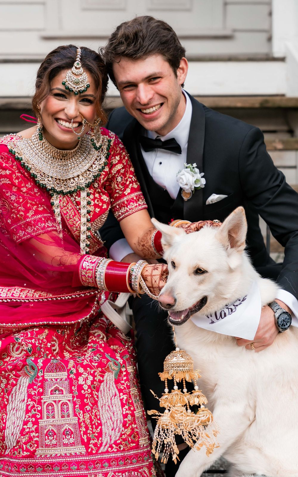 Wedding couple in traditional Indian attire pose joyfully with their white dog wearing a bow tie at their celebration.