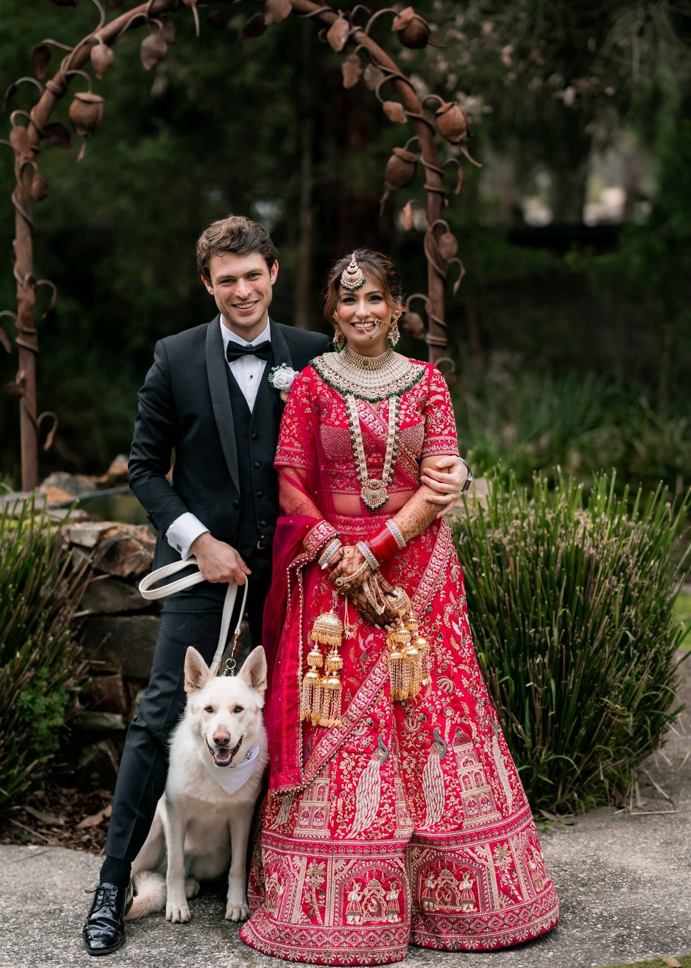 A couple in wedding attire sits outdoors with their white husky dog on a garden path.