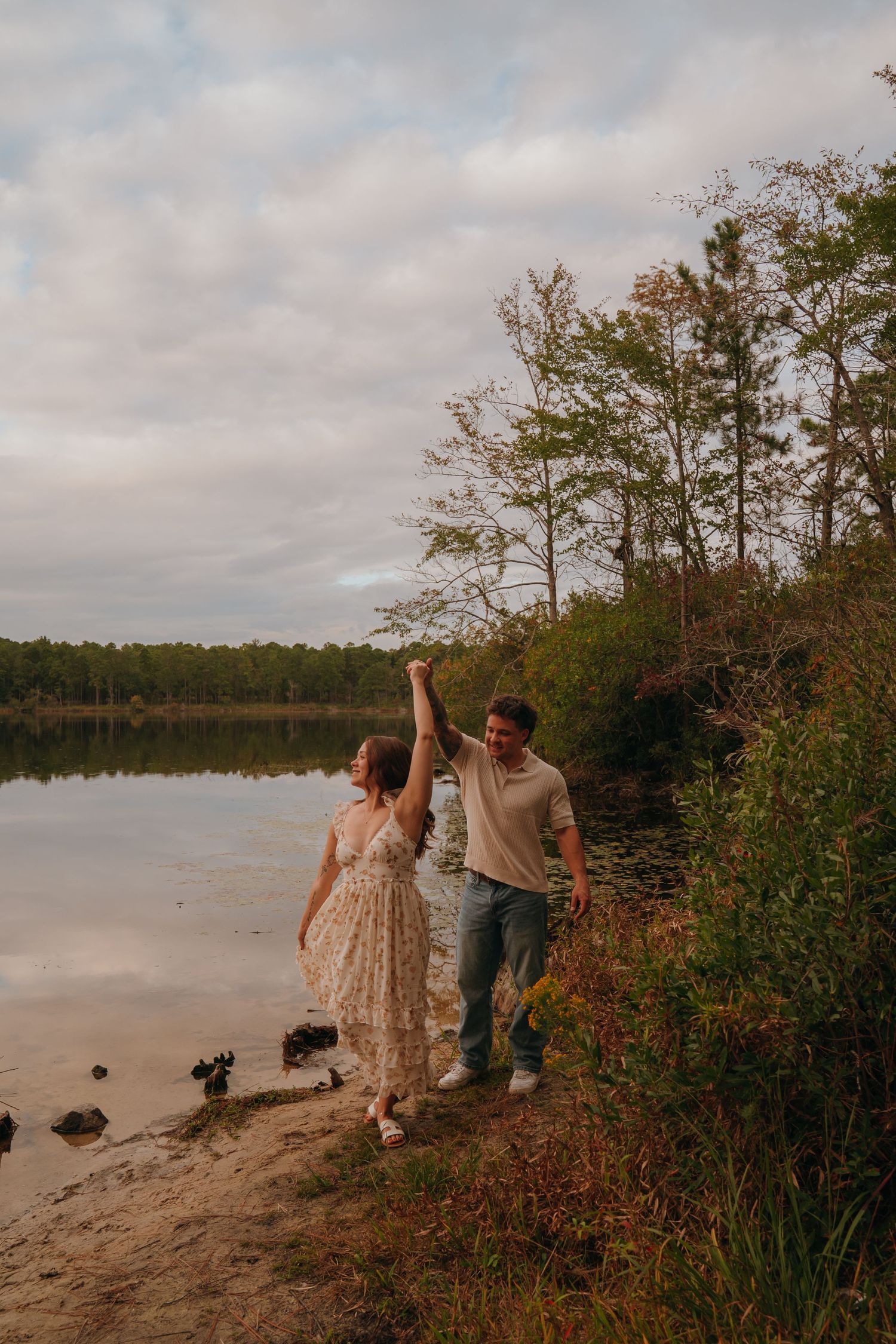 A couple walks hand in hand along a winding brick path under tall trees in a romantic park setting.