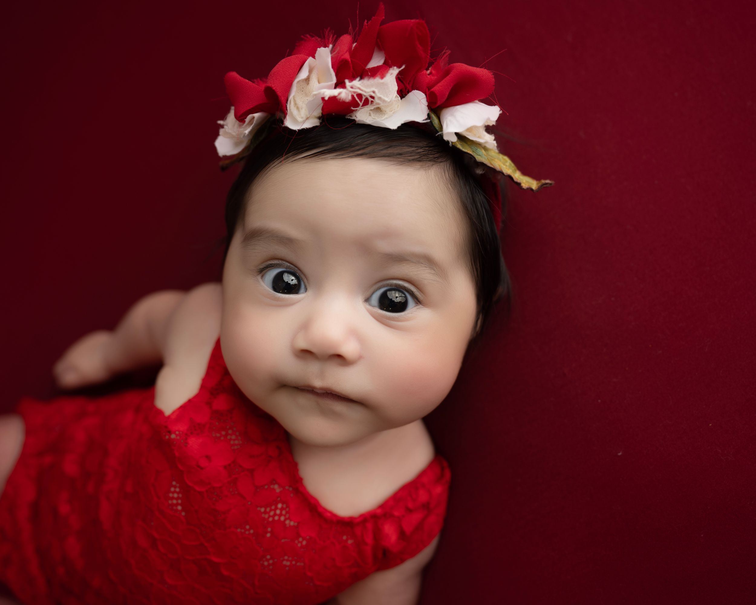 Baby wearing red dress and floral crown against deep red background.