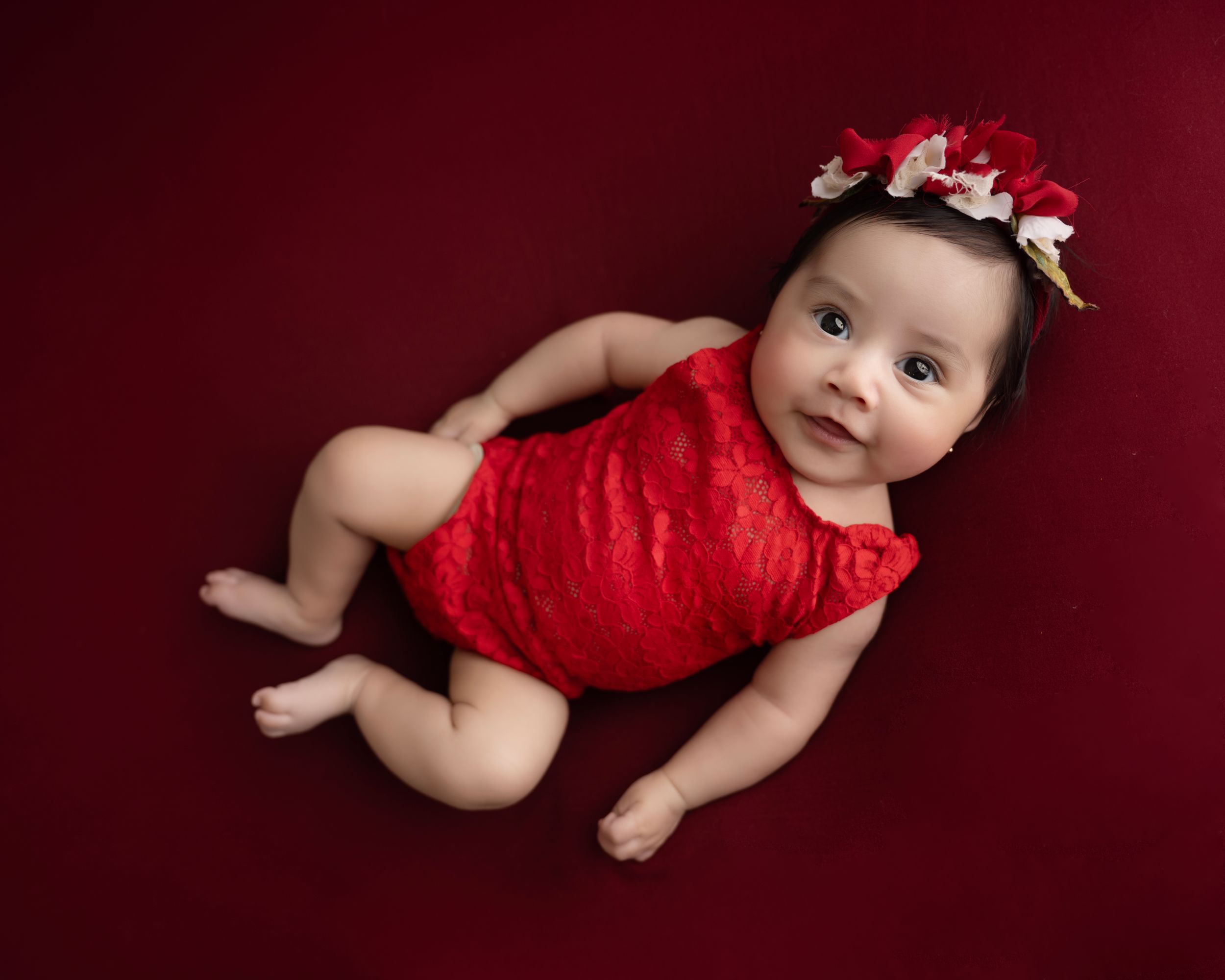 Baby wearing red lace romper and floral headband lying on burgundy background during professional photoshoot.