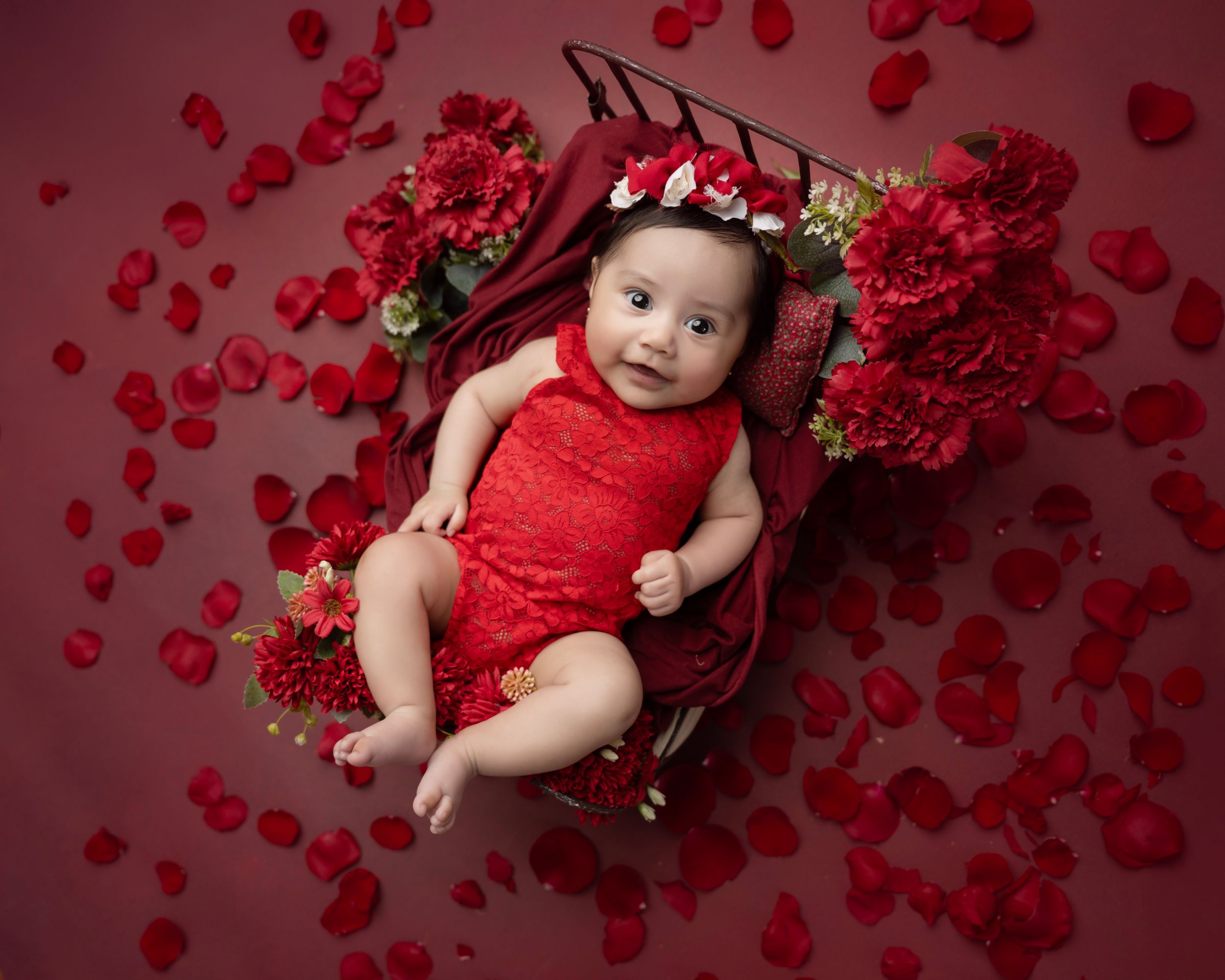 Baby in red outfit lying in wooden basket with scattered rose petals and red flowers.