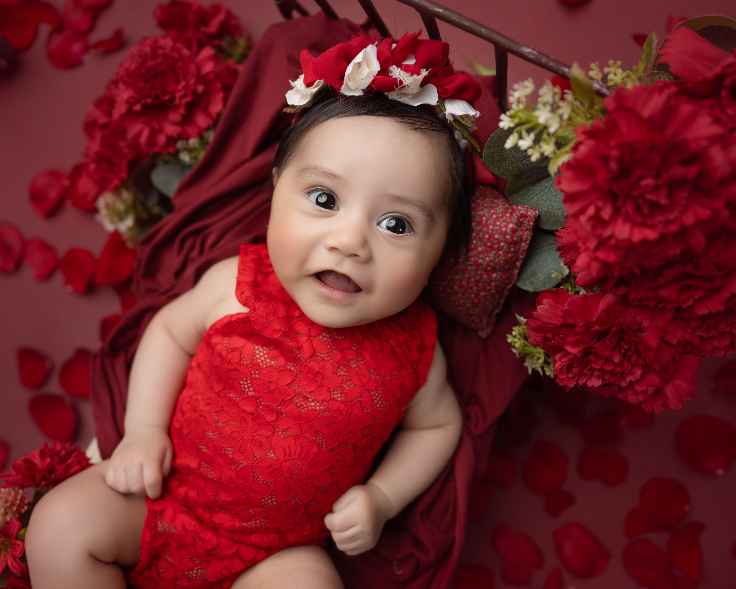 Baby in red outfit surrounded by red carnations and floral decor during studio photography session.