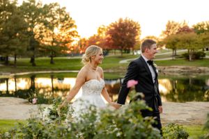 A romantic couple walking hand in hand by a tranquil pond at sunset during an elegant outdoor wedding.