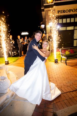 Newlyweds dance together on brick pavement with sparklers creating magical light effects outside Piropos restaurant at night.