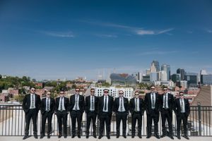 A group in matching black suits and ties stand in a row on a rooftop terrace with the Kansas City skyline behind them.