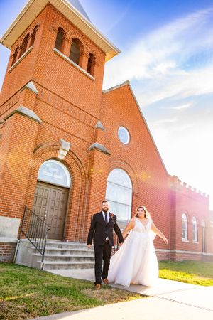 A couple descends the steps of a historic red brick church with arched windows during their wedding celebration.