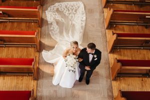 Aerial view of couple in wedding attire sitting between wooden church pews.