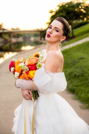 Bride in white off-shoulder dress holds vibrant bouquet of orange, red and yellow flowers at sunset by garden path.