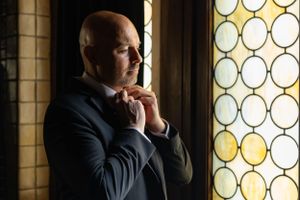 A contemplative person wearing a dark suit stands by an ornate window with circular stained glass patterns.