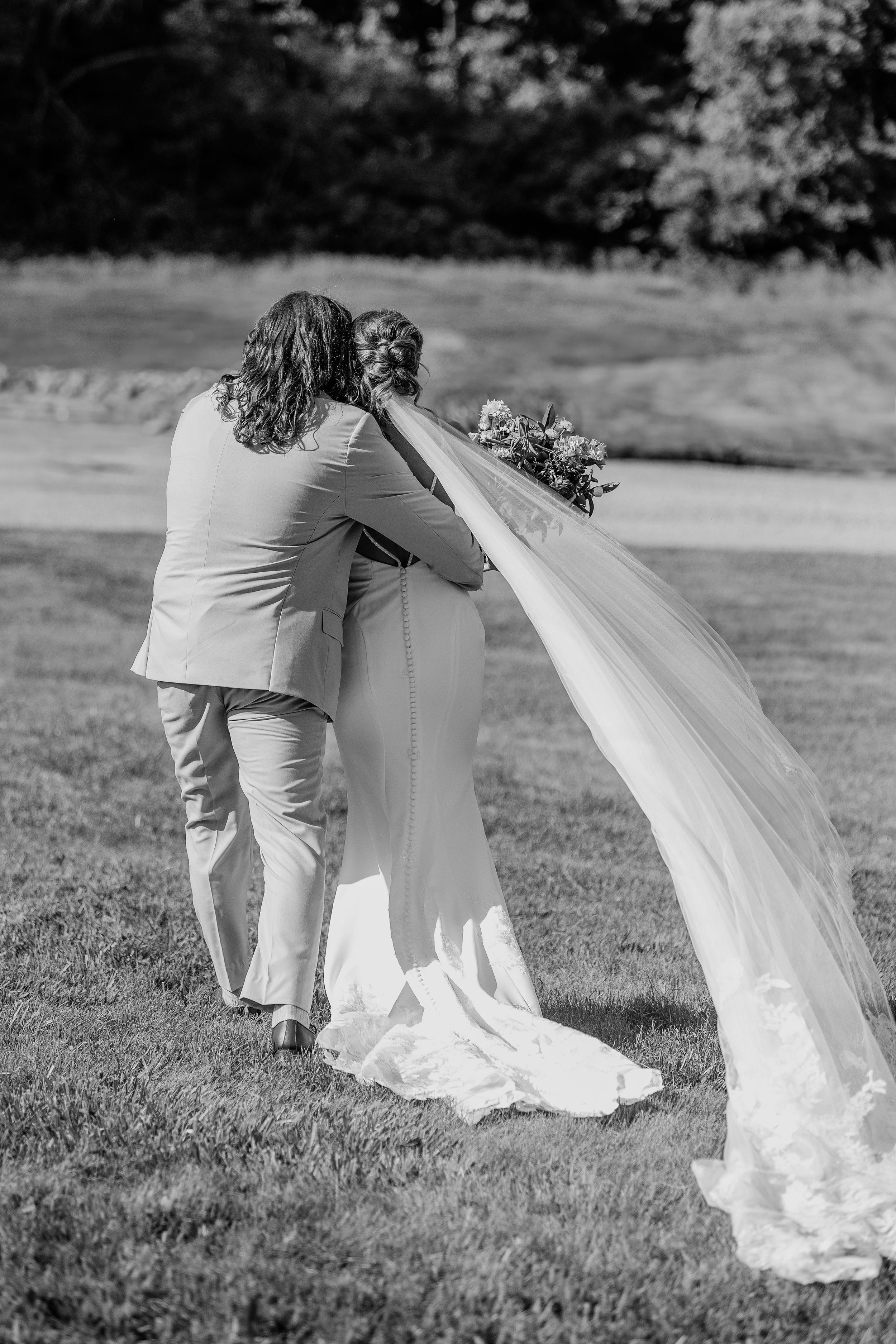 Monochrome photo of wedding couple embracing as veil flows in the wind on grassy lawn.