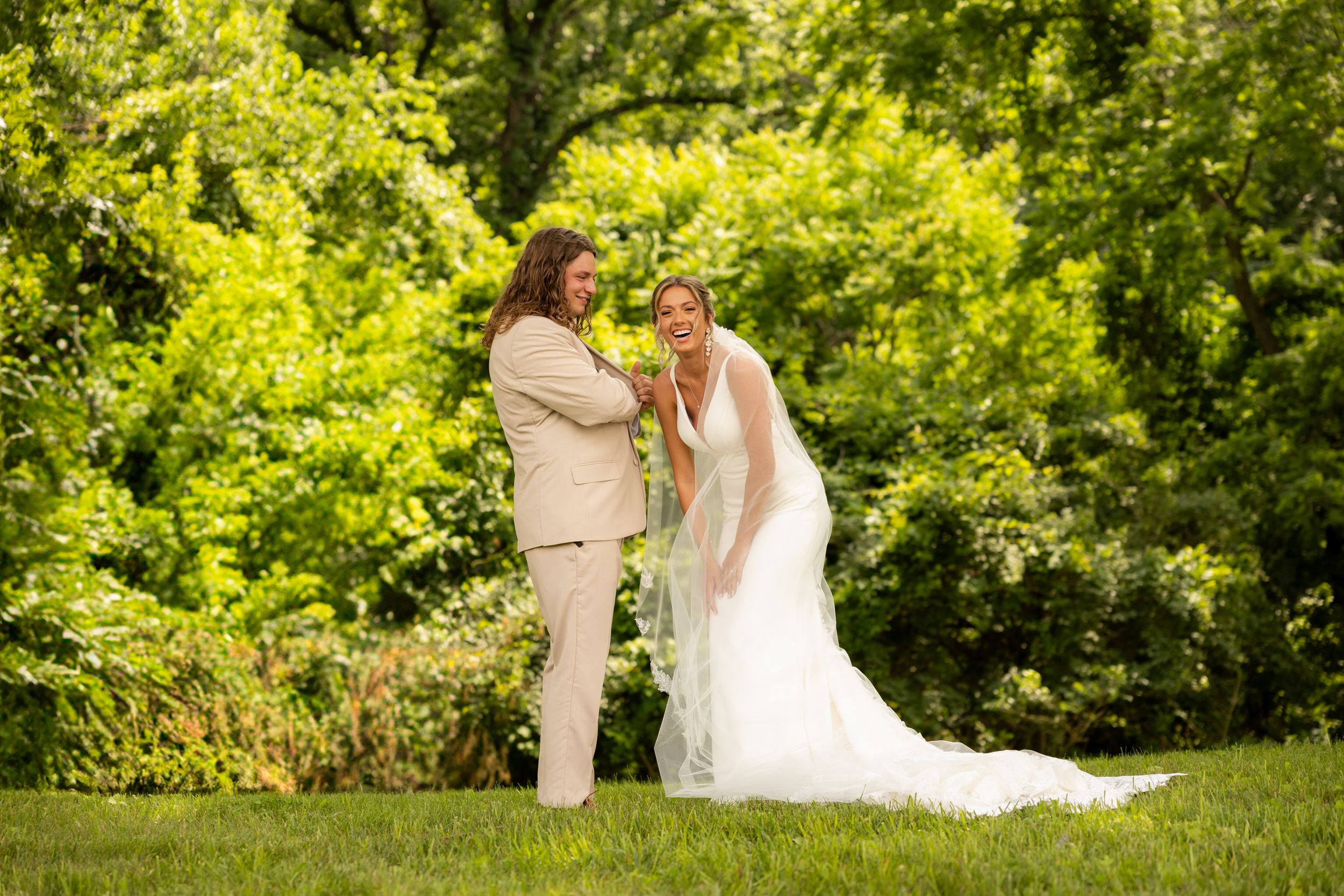 A romantic wedding couple shares a tender moment outdoors in a lush green garden setting on a sunny summer day.