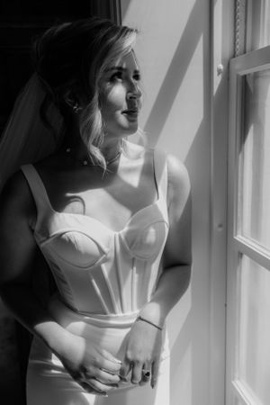 Black and white portrait of a bride in a structured white corset dress standing by a sunlit window.