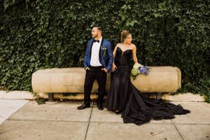 Two elegantly dressed people sit on stone bench against an ivy wall, wearing formal black attire with blue bouquet.