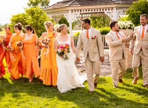 Wedding party in orange bridesmaids dresses and tan suits walk across sunny lawn during outdoor summer celebration.