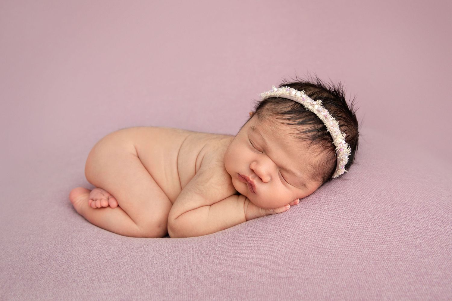 Peaceful sleeping newborn with lace headband on pink blanket in artistic pose.