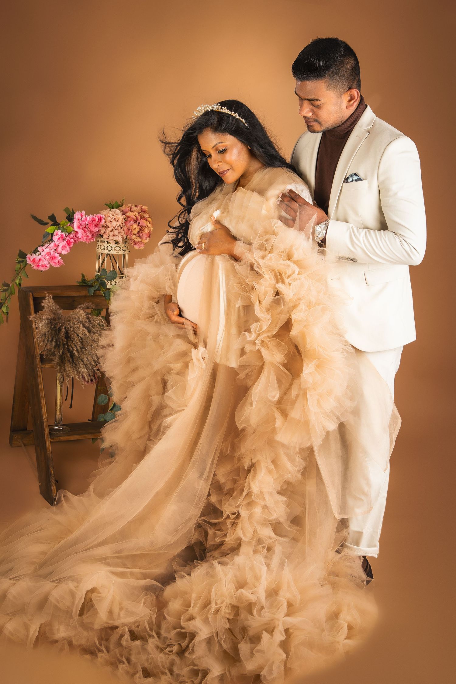 A pregnant woman in a fluffy dress with her partner looking down on her pregnant belly with some flowers at the back