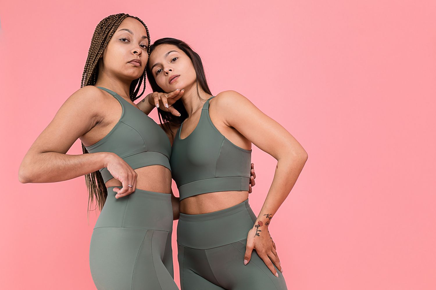 Two models wearing sage green workout sets pose confidently against a pink background.