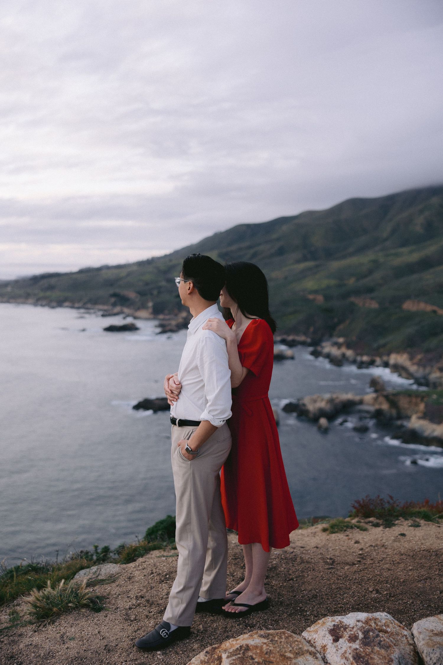 A couple stands on a coastal cliff overlooking the rugged Big Sur coastline in California at dusk.