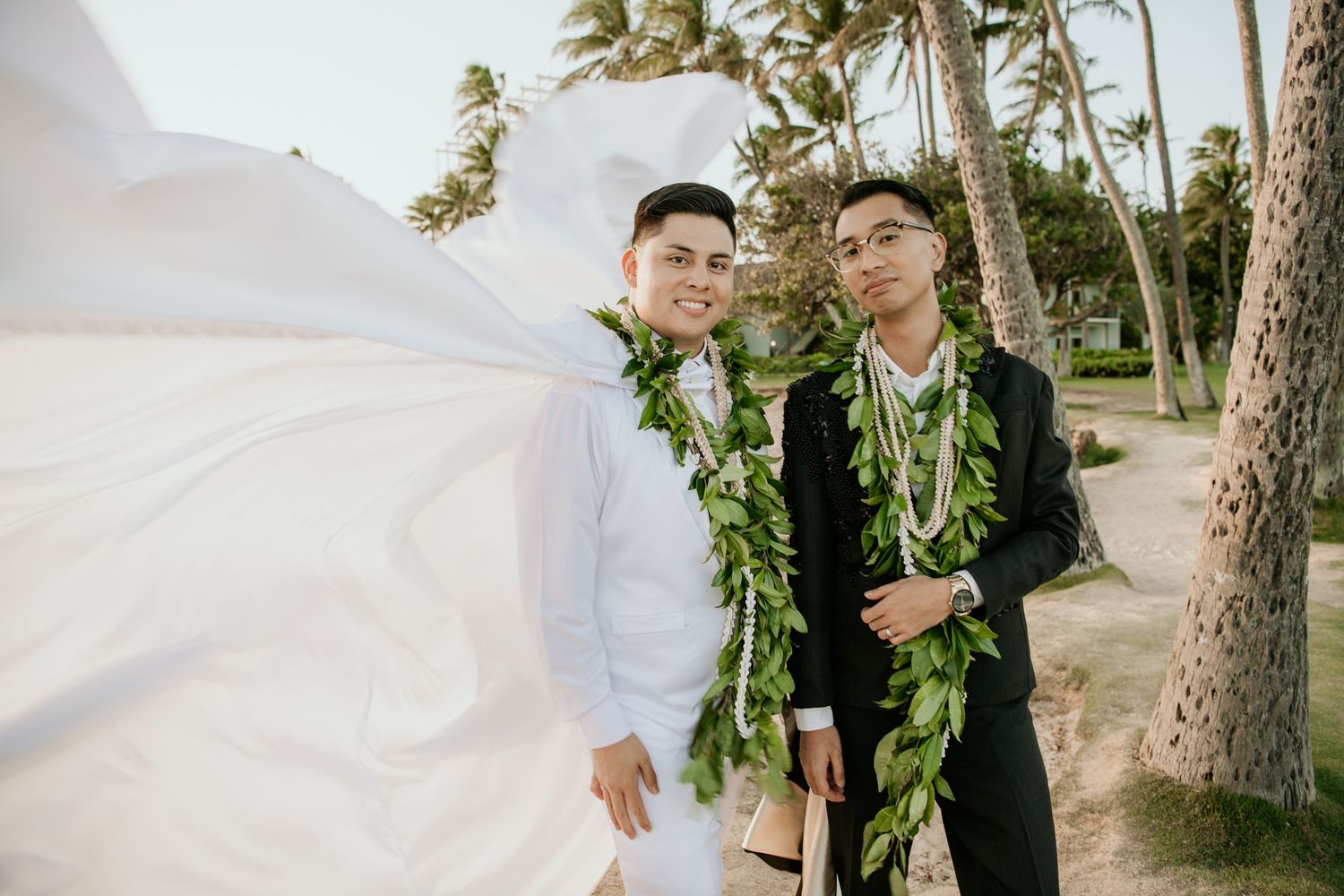 Two people wearing traditional Hawaiian lei garlands stand together on a tropical beach with palm trees in the background at Kahala Hotel.