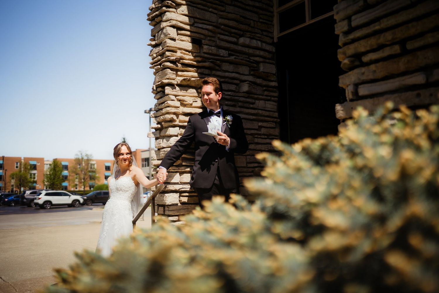 Bride and groom holding hands near a rustic stone building outdoors on a sunny wedding day.
