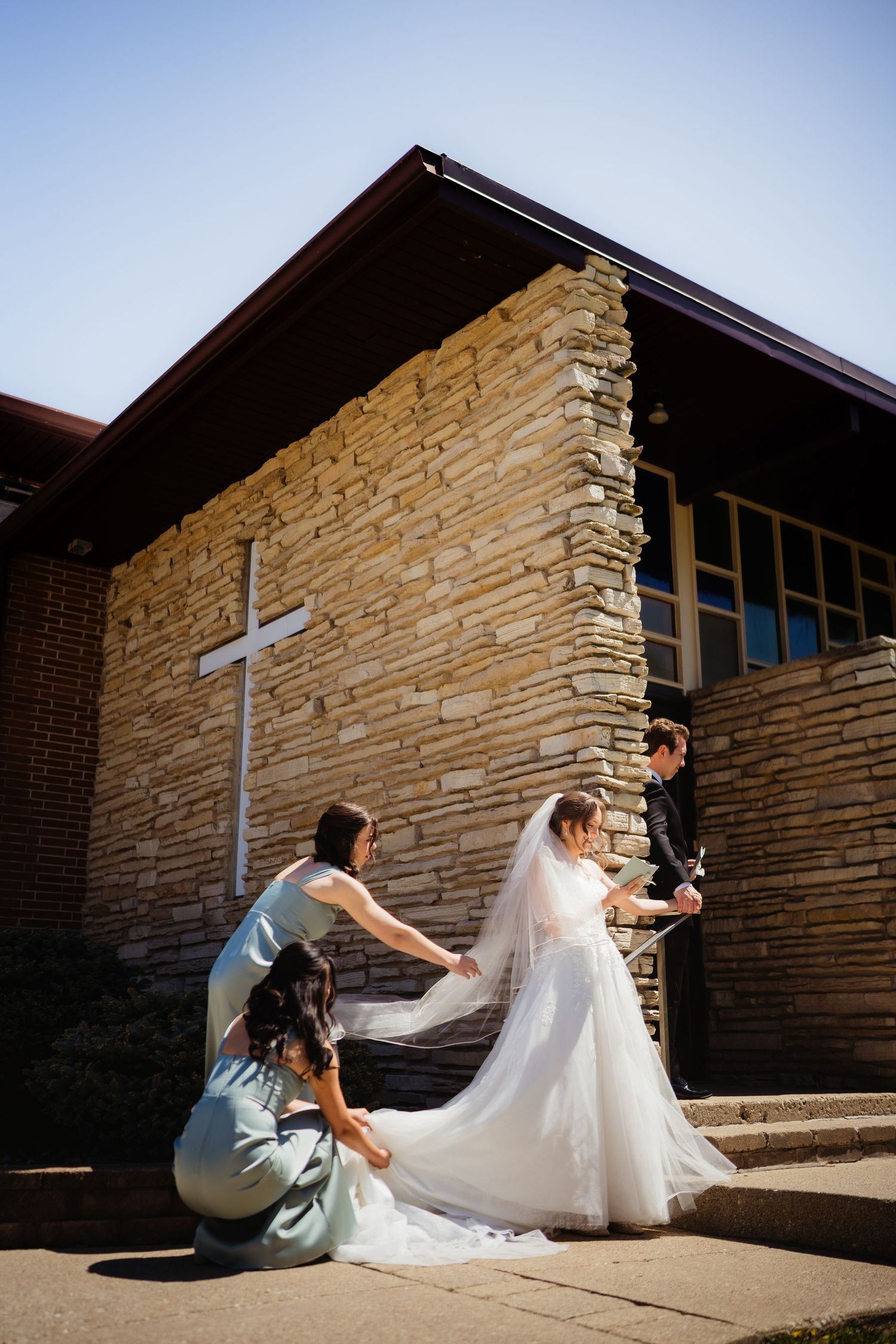 Bride getting her wedding dress arranged by bridesmaids outside a stone church on a sunny day.