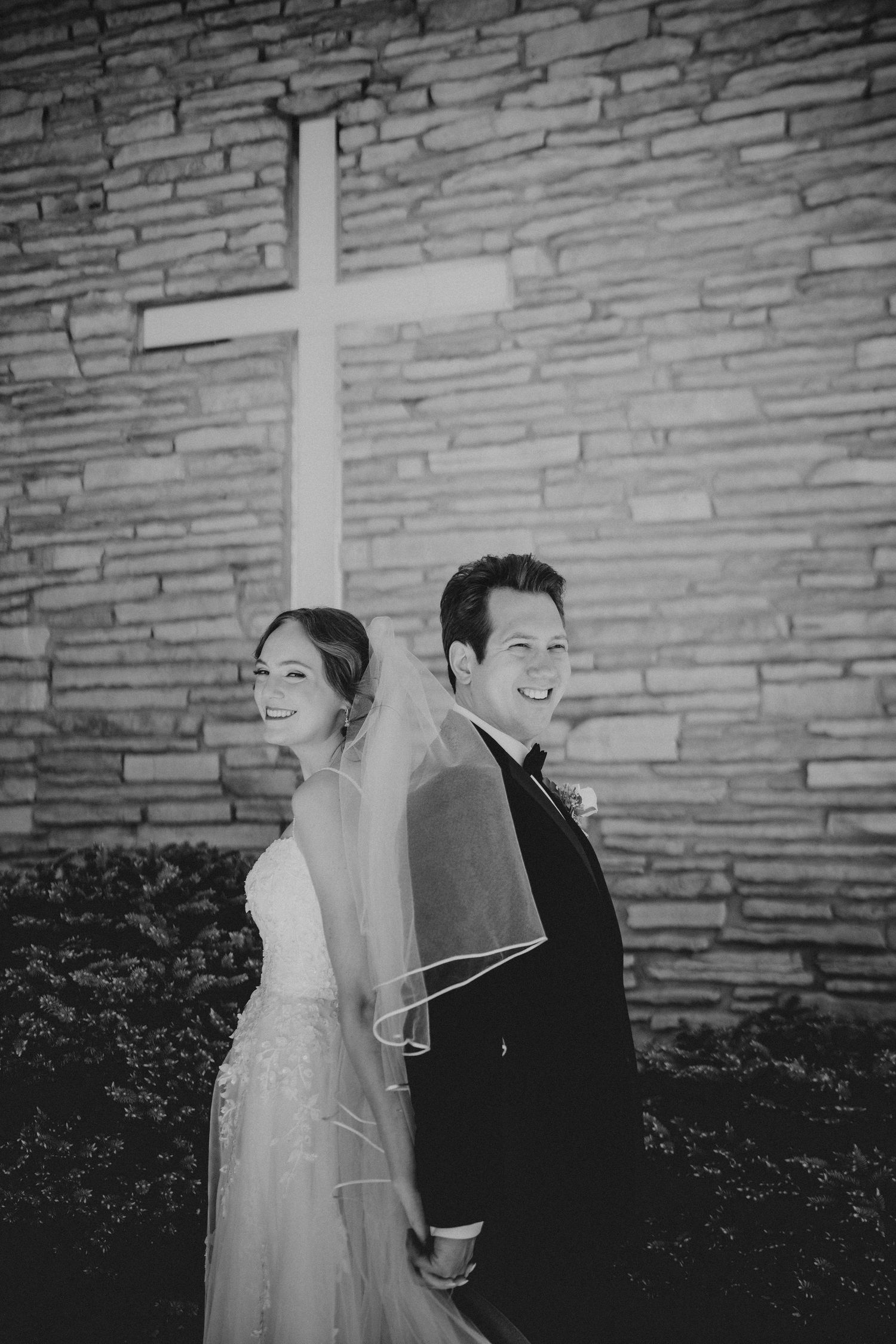 Black and white photo of smiling bride and groom standing back-to-back before a stone church wall with a white cross.