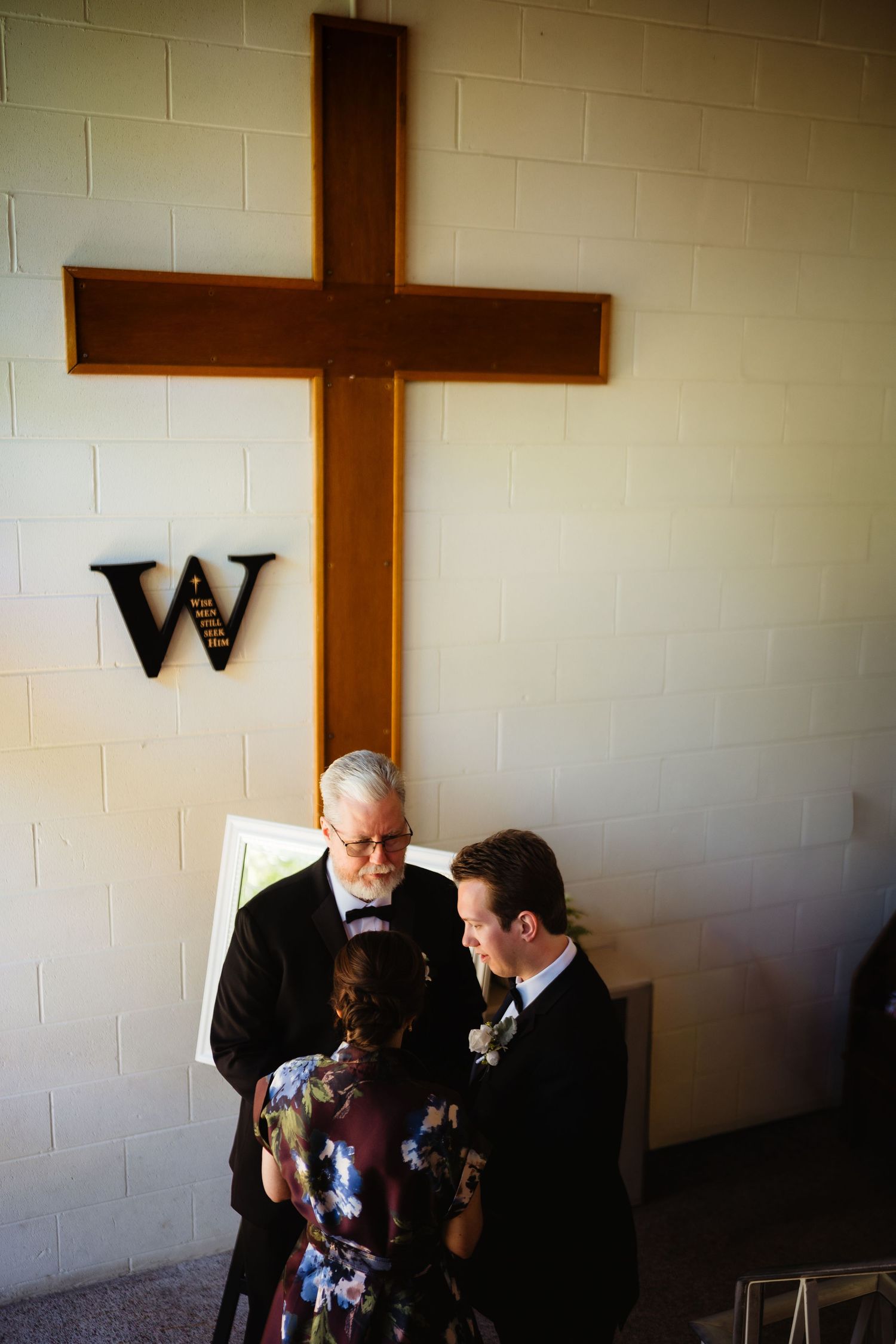 Couple exchanging vows before officiant under wooden cross in church, wedding ceremony with floral dress and boutonniere.