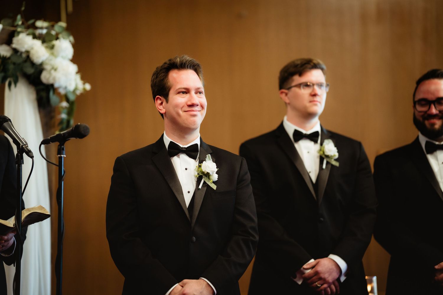 Groom smiling at altar in tuxedo with boutonnière, flanked by groomsmen in matching black suits at wedding ceremony.