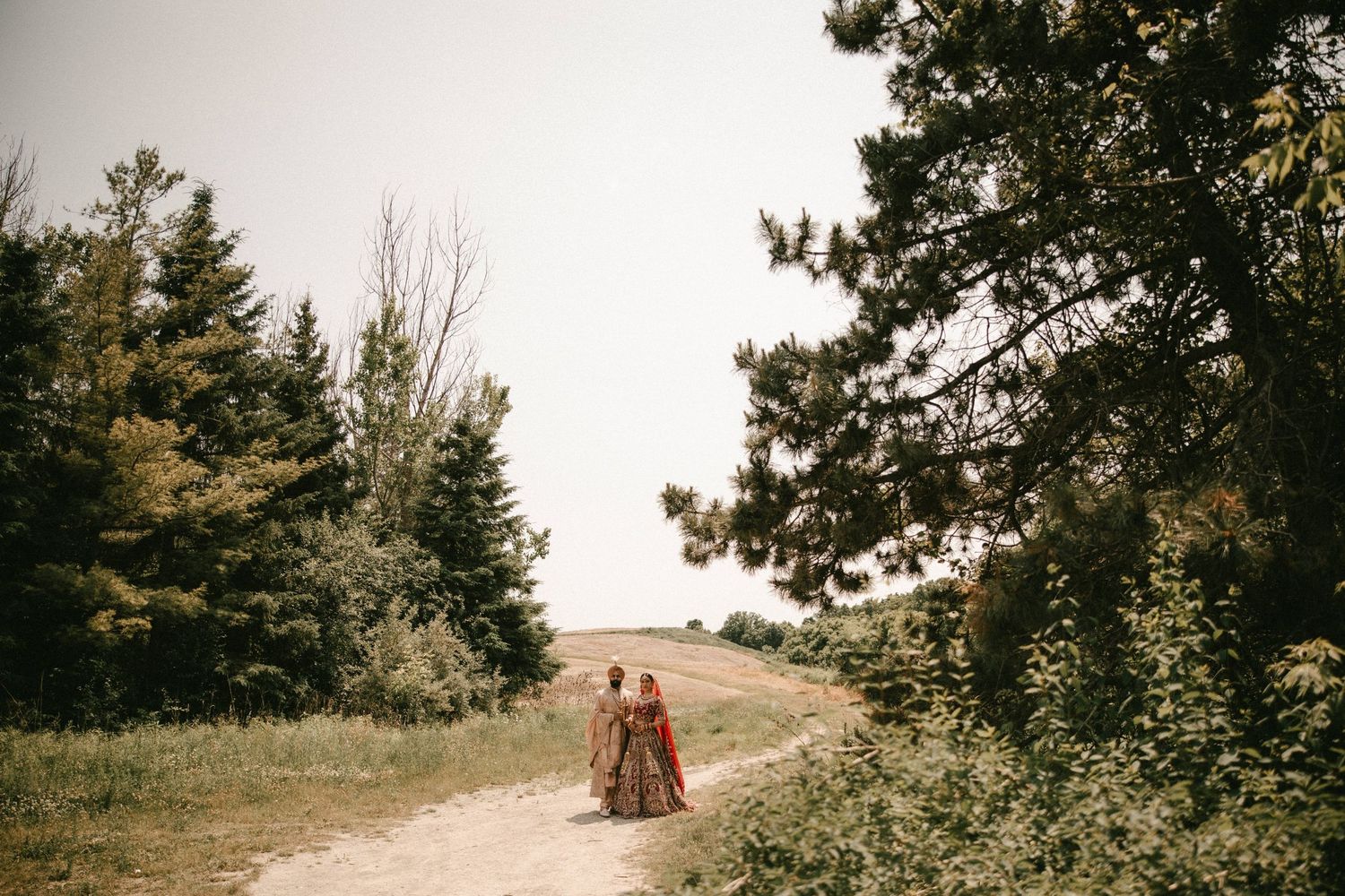 Couple in colorful traditional attire walking hand in hand on a scenic dirt path surrounded by lush green trees.