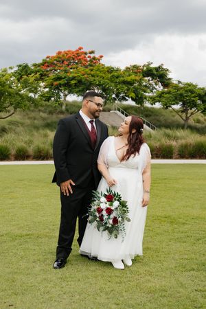 Bride and groom smiling at each other on green lawn, she holds red rose bouquet, he wears black suit with burgundy tie.