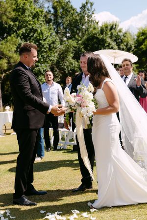 Bride and groom exchanging vows at outdoor wedding ceremony on green lawn with guests and trees in background.