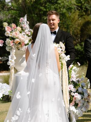 Bride in white gown and floral veil faces groom in black tuxedo at outdoor wedding ceremony altar with pink flowers.