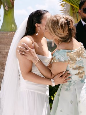 Bride in white gown and veil shares a kiss with a guest in a floral dress at an outdoor wedding ceremony.