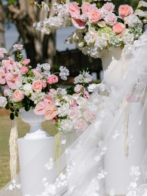 Elegant wedding arch adorned with pink roses, ranunculus, and white florals beside a white urn vase outdoors.