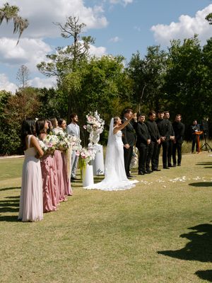 Wedding party stands outdoors as bride and groom share first kiss, bridesmaids in pink and groomsmen in black suits.