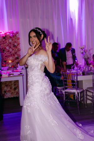 Bride in white lace mermaid gown making peace signs on dance floor at elegant pink-lit wedding reception.