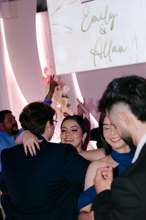 Guests celebrate and embrace at Emily and Allan's wedding reception under a glowing sign.