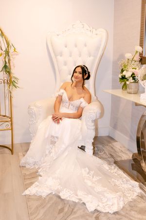 Bride in white lace mermaid wedding dress seated on ornate tufted white throne chair in bridal suite.