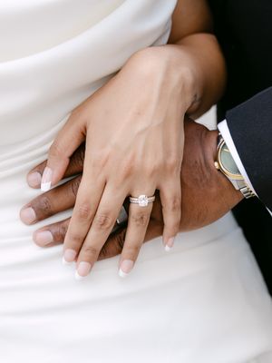 Close-up of a bride and groom's hands showing a diamond engagement ring and wedding band, with the groom wearing a gold watch.