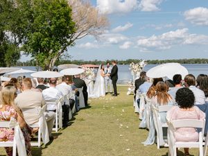 Outdoor lakeside wedding ceremony with guests holding white umbrellas as couple exchanges vows at floral arch.