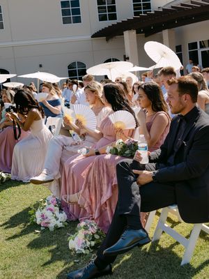 Wedding guests in pink bridesmaid dresses holding fans and parasols at an outdoor ceremony on a sunny day.