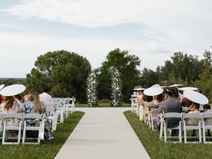 Outdoor wedding ceremony with guests seated in white chairs along a grass aisle leading to a floral arch.