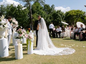 Outdoor wedding ceremony on green lawn with bride in long veil, groom, floral arrangements, and seated guests under trees.
