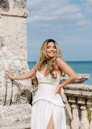 Woman in white corset dress smiling by stone balustrade with ocean backdrop.
