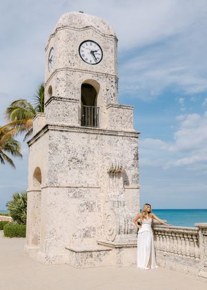 Bride in white gown poses by historic Palm Beach clock tower with ocean backdrop and palm trees.